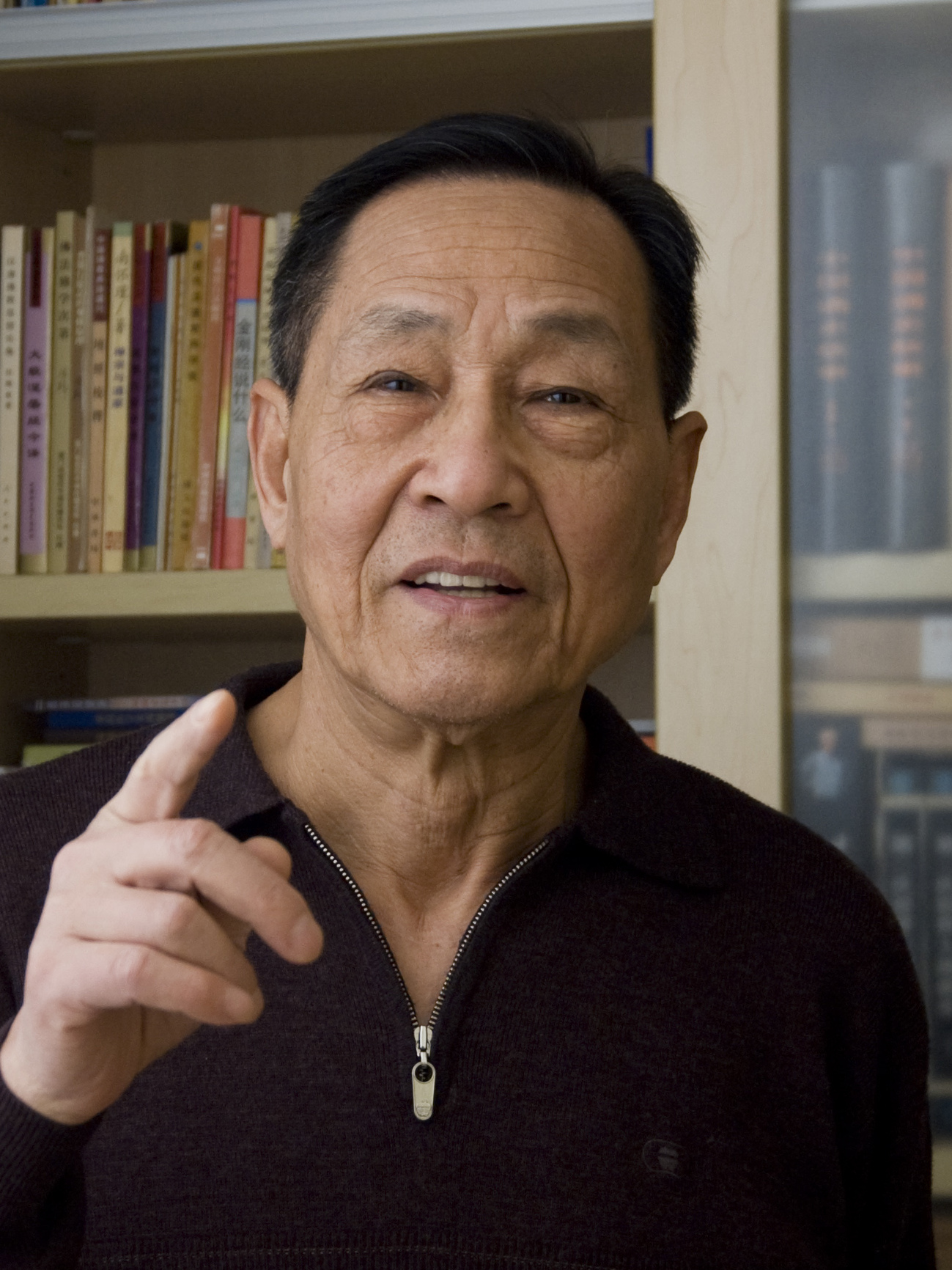 Bao Tong sitting before bookshelves with one hand raised.