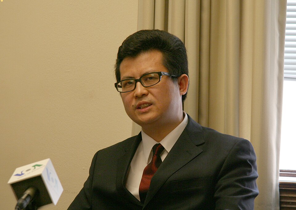 Guo Feixiong in a suit and glasses with microphones in front of him.