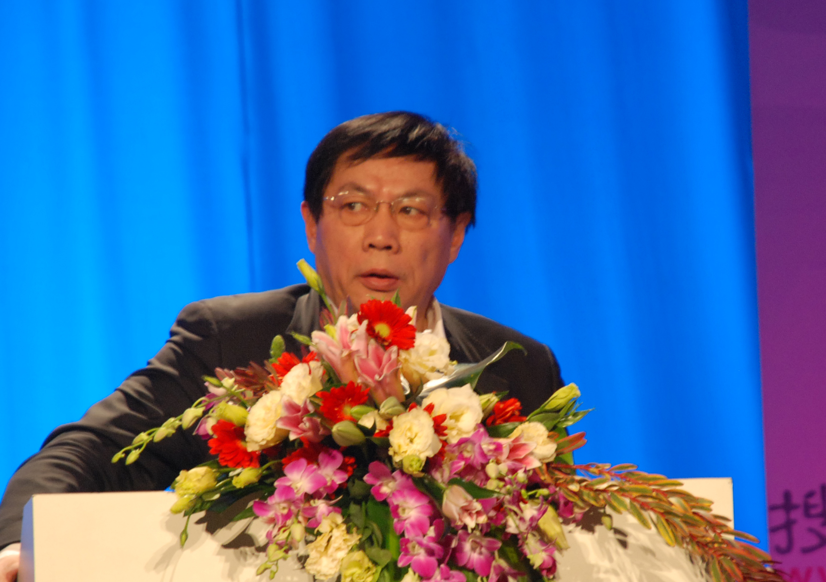 Ren Zhiqiang speaking behind a lectern decorated with flowers, against a blue curtain.