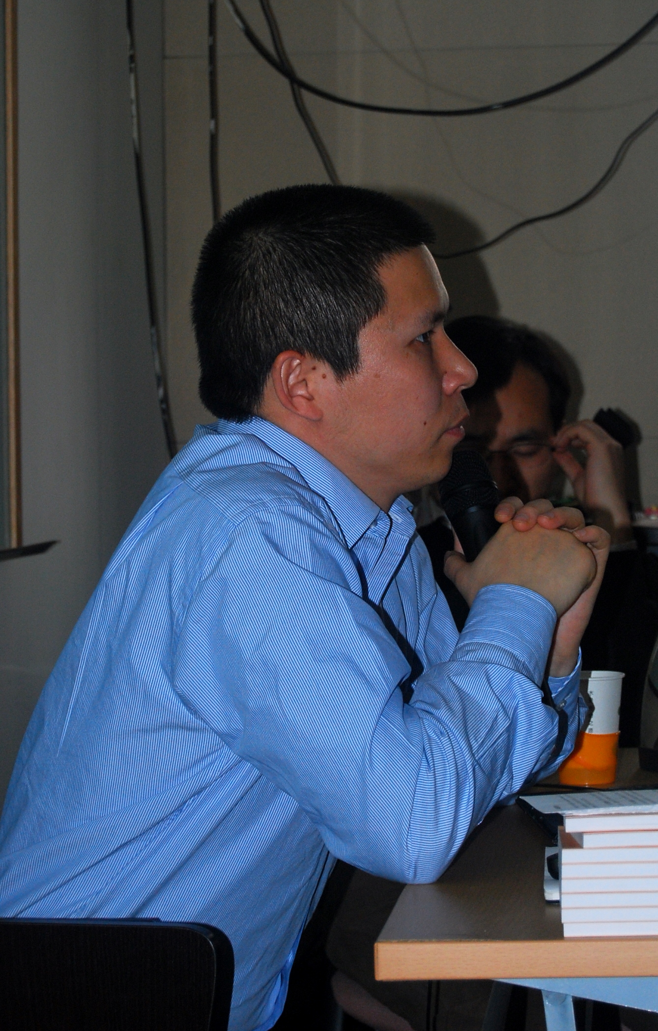 Xu Zhiyong wearing a blue striped shirt, seated sideways at a table holding a microphone.